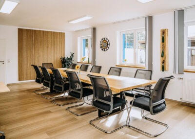 Ein großer Konferenztisch aus Holz steht in einem modernen Besprechungsraum, umgeben von schwarzen Stühlen; Details wie Fenster und Pflanzen verleihen dem Raum Helligkeit.