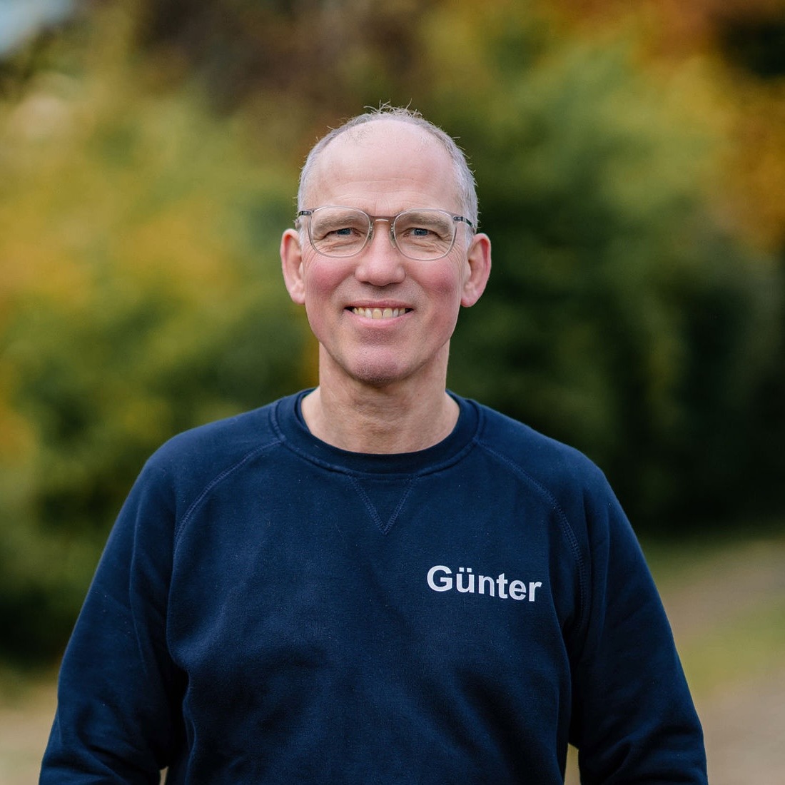 Ein Mann mit Brille und blauer Sweatshirt steht lächelnd im Freien vor grünem Hintergrund. Sein Sweatshirt trägt den Namen ''Günter''.