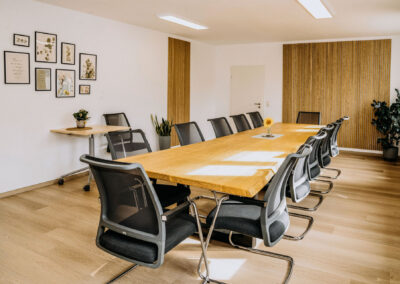 Ein moderner Besprechungsraum mit großem Holzkonferenztisch und ergonomischen Stühlen. An der Wand hängen Bilder mit Pflanzenmotiven.