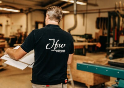 Eine Person mit T-Shirt, auf dem ''Jfw'' steht, trägt mehrere Papiere in einer Werkstatt. Der Raum ist hell erleuchtet und zeigt verschiedene Werkzeuge.
