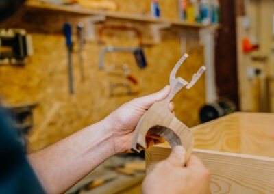 Eine Person hält ein handgefertigtes Holzteil in der Werkstatt, umgeben von Werkzeugen und Materialien. Das Holzstück hat eine komplexe Form.