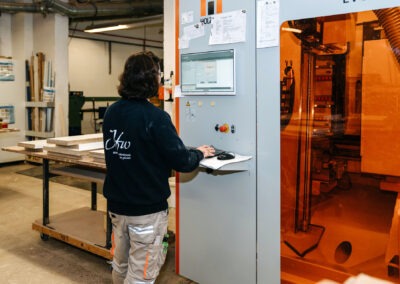 Eine Person steht vor einer digitalen Steuerung in einer Holzwerkstatt und bedient eine Maschine zur Bearbeitung von Holzmaterial.