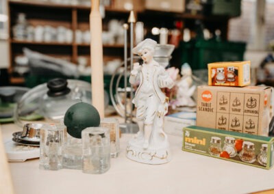Auf einem Tisch ist eine Sammlung von verschiedenen Haushaltsgegenständen zu sehen, darunter eine Porzellanfigur, Kerzen und Streuer; die Gegenstände sind vielfältig angeordnet.