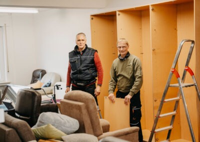 Zwei Männer stehen in einem Raum mit Möbeln und einem offenen Schrank. Der Mann links trägt eine dunkelblaue Weste, der Rechts einen grünen Pullover und hält eine Holplatte.