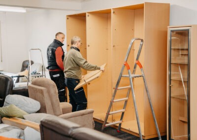 Zwei Männer bauen Möbel zusammen; einer hält eine Holzplatte, während der andere im Hintergrund lächelt. Eine Leiter steht neben dem Möbelstück in einem Raum mit Sitzmöbeln.