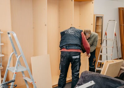 Zwei Personen installieren einen Korpus für ein Möbelstück in einem Raum mit hellen Wänden. Eine Person hält ein Bauteil, während die andere darauf achtet.