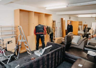 Drei Personen arbeiten in einem Raum voller Möbel und Regale; sie montieren zusammen einen Schrank, während sie umgebauten Plätzen nutzen.