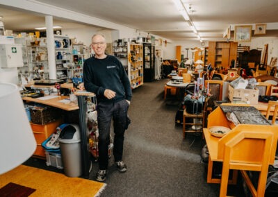 Ein Mann mit Brille und schwarzem Pullover lächelt im Innenraum eines Second-Hand-Ladens, umgeben von Regalen mit Haushaltswaren und Möbeln.