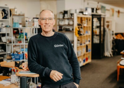 Ein Mann steht in einem Verkaufsgeschäft mit einem schwarzen Sweatshirt und Brille; im Hintergrund sind Regale mit verschiedenen Haushaltswaren sichtbar.