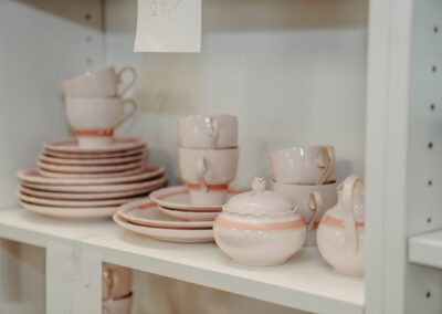 Teller und Tassen in sanften Rosa- und Beigetönen auf einem Regal. Die Geschirrteile sind kunstvoll verziert und sorgfältig angeordnet.