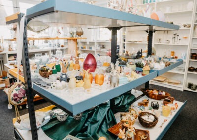 Ein bunter Tisch mit verschiedenen Dekorationsgegenständen und Souvenirs, darunter Eier, Hasenfiguren und Gläser, in einem hellen Raum auf regalen.