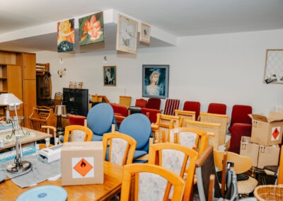 Ein vollgestellter Raum mit Möbeln, Kartons und Bildern an den Wänden; Stühle und Tische sind unordentlich angeordnet, mit einer Gefahrgutbox auf einem Tisch.
