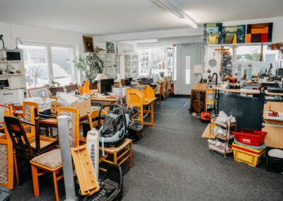Ein Ausstellungsraum mit verschiedenen Möbelstücken und Haushaltsgegenständen; Stühle stehen übereinander, während Tische und Regale eine Vielzahl an Artikeln präsentieren.