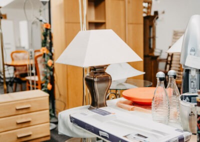 Eine schöne Tischlampe auf einem Tisch neben mehreren Gegenständen wie Wasserflaschen, einer Schüssel und einem Karton, vor einem hellen Hintergrund.