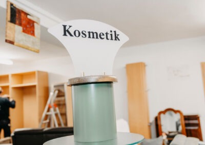 Ein Schild mit der Aufschrift ''Kosmetik'' steht auf einem transparenten Stand, im Hintergrund ist ein Möbelstück zu sehen.