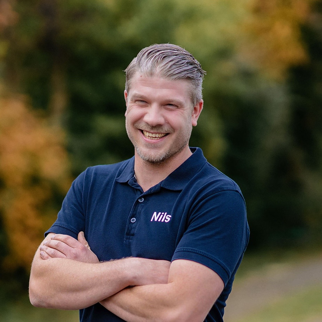 Ein lächelnder Mann mit kurzen, blonden Haaren steht im Freien mit verschränkten Armen. Er trägt ein dunkelblaues Poloshirt mit dem Namen ''Nils'' in rosa Schrift.