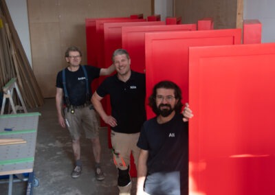 Drei Männer stehen in einem Raum mit roten Türen, lächelnd und in Arbeitskleidung. Im Hintergrund sind Holzplatten und Werkzeuge zu sehen.
