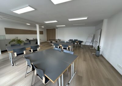 Moderner Seminarraum mit hellen Wänden, Tischen und Stühlen. Der Raum ist gut beleuchtet und bietet Platz für Gruppenaktivitäten, ideal für Schulungen oder Besprechungen.
