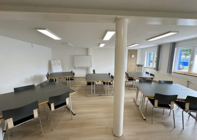 Ein helles und modernes Schulungsraum mit mehreren schwarzen Tischen und Stühlen, die ordentlich angeordnet sind. In der Ecke steht eine Tafel.