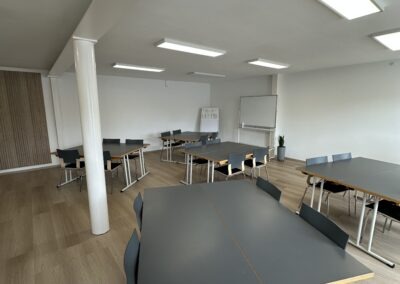 Ein moderner Seminarraum mit mehreren rechteckigen Tischen und Stühlen in hellem, neutralem Farbdesign, ideal für Schulungen oder Besprechungen.
