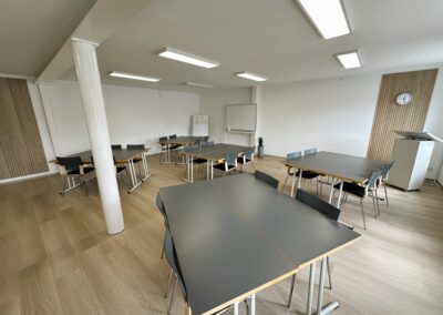 Moderne Unterrichtsräume mit grauen Tischen und Stühlen in einem hellen Raum; eine Uhr und ein Flipchart sind sichtbar.