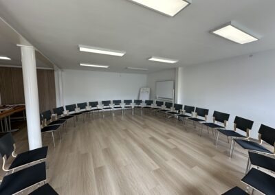 Ein heller, moderner Seminarraum mit einem runden Sitzkreis aus schwarzen Stühlen und einem Holzfußboden. Die Wände sind schlicht und weiß, mit einer Tafel im Hintergrund.