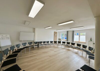 Ein heller Raum mit einer kreisförmigen Anordnung von schwarzen Stühlen; im Hintergrund steht eine Tafel mit dem Schriftzug "Herzlich Willkommen".