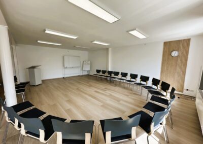 Ein leerer Seminarraum mit kreisförmig angeordneten Stühlen und einem Whiteboard in heller Umgebung. Die Holzoptik des Fußbodens sorgt für eine freundliche Atmosphäre.