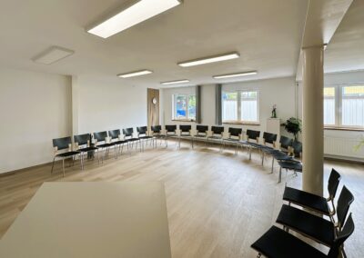 Ein heller Seminarraum mit Stühlingsarrangement; in einem großen, modernen Raum stehen Stühle in einem Halbkreis um einen Tisch, es gibt große Fenster und eine Uhr an der Wand.