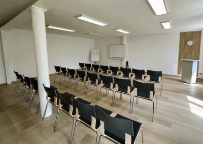 Ein gut beleuchteter Seminarraum mit Reihen von schwarzen Stühlen und einem Rednerpult; die Wände sind schlicht und hell gehalten.