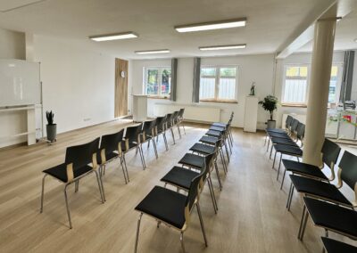 Ein heller Seminarraum mit mehreren Stuhlreihen, einer Leinwand und einem Pult; der Raum ist ordentlich und gut beleuchtet.