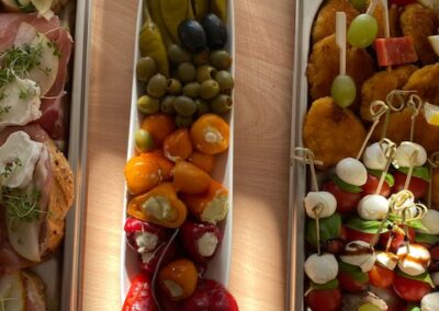 Ein gedeckter Tisch mit verschiedenen Vorspeisen. In der Mitte ist eine lange Schale mit buntem Gemüse, darunter Paprika und Oliven, umgeben von weiteren Platten mit Snacks.