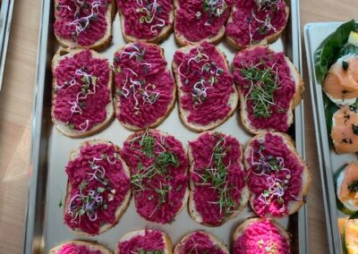 Eine Silberschale mit belegten Brötchen, die mit einer lila Paste und frischen Kräutern dekoriert sind. Die Brötchen sind gleichmäßig angeordnet und wirken appetitlich.