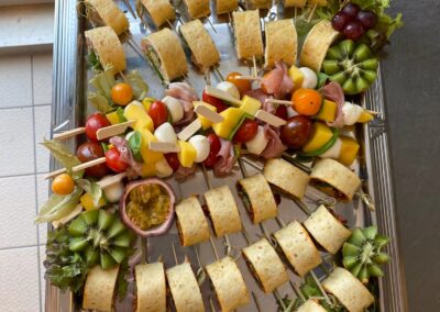 Ein Buffet mit verschiedenen kleinen Snacks, darunter gefüllte Wraps, Gemüse-Spieße und frische Früchte wie Trauben und Kiwi auf einer silbernen Platte.