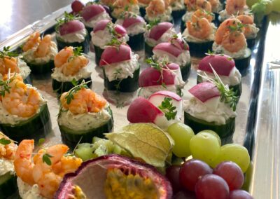 Ein platter mit eleganten Häppchen, bestehend aus Garnelen, Frischkäse und Gurke, dazu frische Trauben und Passionsfrucht.