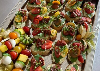 Eine Platte mit diversen belegten Broten, die mit frischen Tomaten, Rucola und getrockneten Tomaten verziert sind, daneben bunte Obstspieße.
