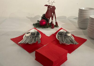 Dezente Tischdekoration mit rotem Besteck und Servietten. Ein stilisierter Elch aus Holz steht neben einem Christbaumkugel und einer grünen Zweigdekoration auf dem Tisch.