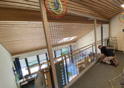 Ein Handwerker installiert ein Sicherheitsgitter im Innenbereich mit Holzdecke und grüner Teppichfläche. Die Gitterstruktur schützt die obere Ebene des Treppenhauses.