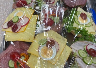 Eine Platte mit belegten Brötchen, garniert mit frischem Gemüse und Fruchtstücken, abgedeckt mit Folie.