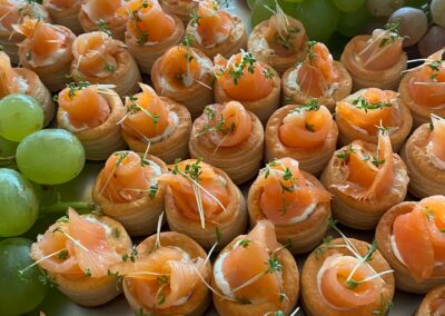 Eine Auswahl an kleinen Häppchen mit Räucherlachs in Teigschalen, umgeben von Trauben und anderen Snacks auf einem Tablett.